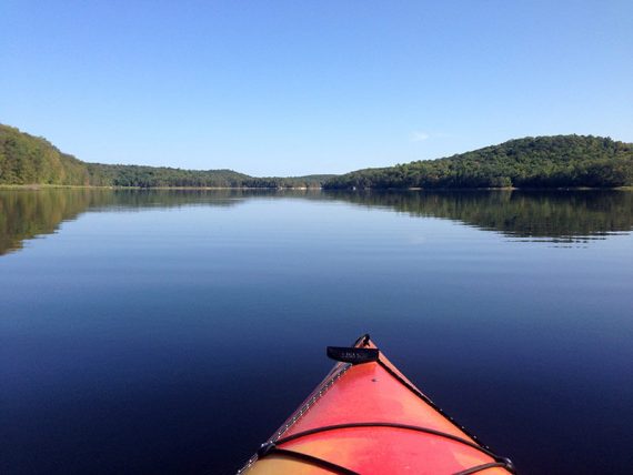 Nose of a kayak seen against calm blue water with green hills beyond.