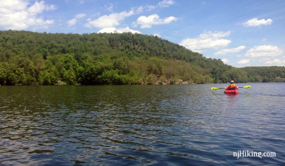 In the background, a tall hill rises behind a kayaker paddling Monksville Reservoir