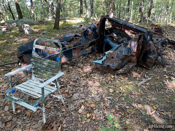 Crumpled and rusted remains of a car next to a chair along a trail