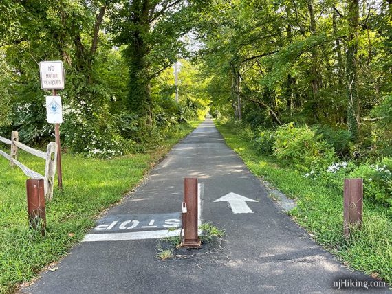 Paved rail trail with metal barrier posts at a crossing