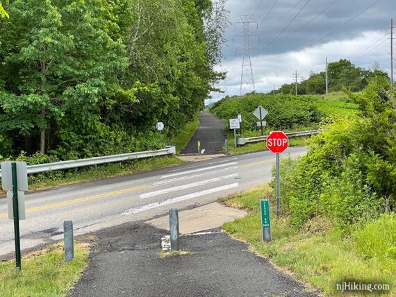 Rail trail at a road crossing with trees on one side and power lines on the other.